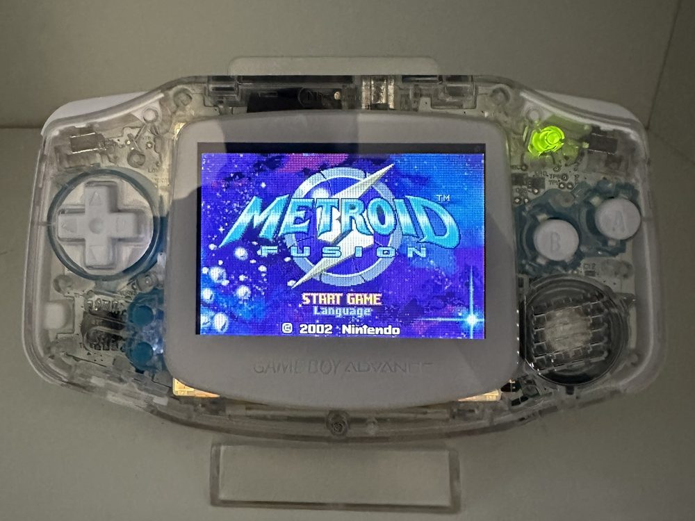 Transparante Game Boy Advance met een helder IPS-scherm dat het titelscherm van Metroid Fusion toont, op een standaard