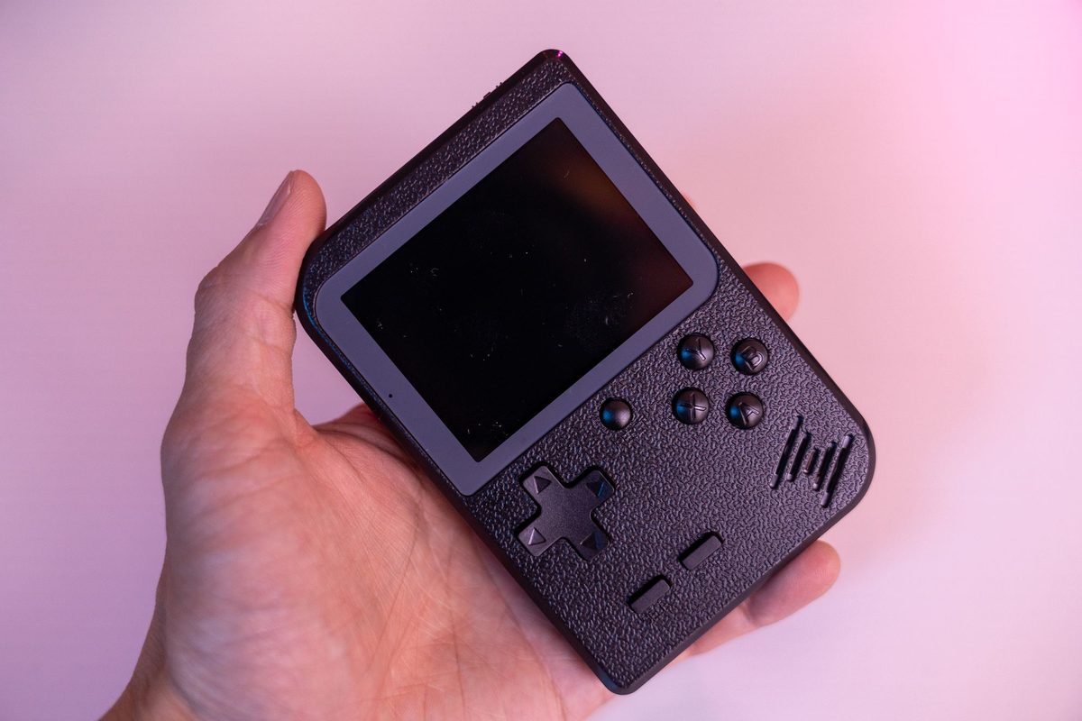 Hand houdt een zwarte retro handheld in Game Boy-stijl met D-pad en vier knoppen vast, tegen een roze achtergrond